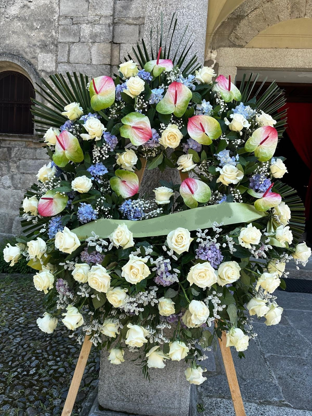 Corona funebre elegante con rose e anthurium freschi, personalizzabile nei colori e fiori, disponibile in misura media (80 cm) e grande (140 cm), con nastro su richiesta.