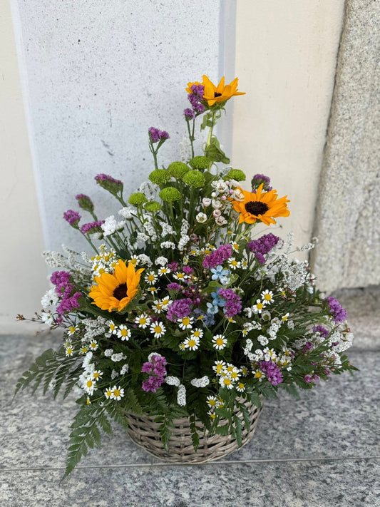Composizione floreale in cesto intrecciato con fiori freschi dai toni campestri giallo, viola, bianco e verde, misura grande.